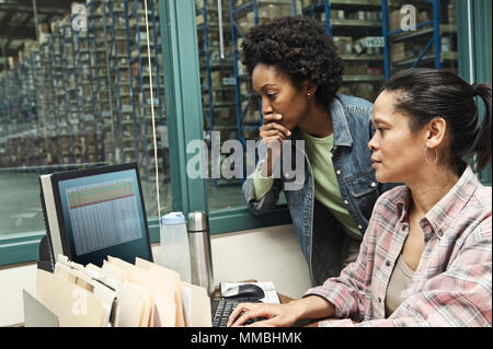 Team of two African American female warehouse workers working on a computer in an office in the middle of a large distribution warehouse full of racks Stock Photo