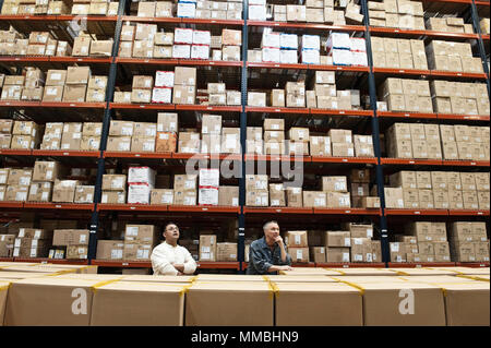 Two male warehouse workers checking inventory in a large distrubiton warehouse full of products stored in cardboard boxes on pallets on large racks. Stock Photo