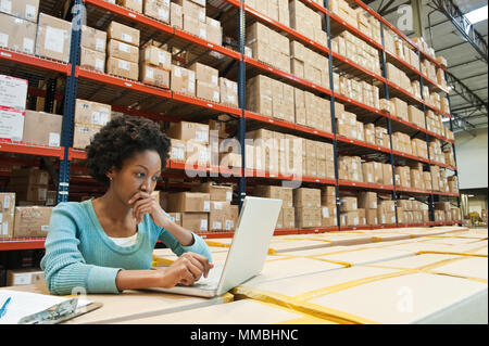 African American female warehouse worker working on inentory on a lap top computer in a large distribution warehouse of products stored in cardboard b Stock Photo