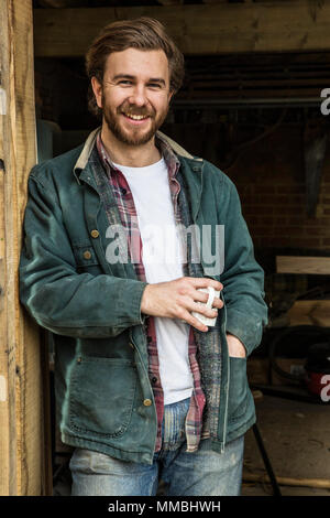 Bearded man standing in doorway of woodworking workshop, holding mug, smiling at camera. Stock Photo