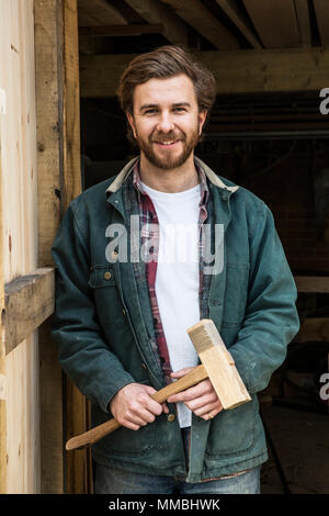 Bearded man standing in doorway of woodworking workshop, holding wooden mallet, smiling at camera. Stock Photo