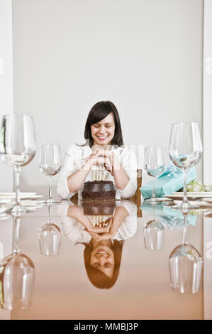 A woman seated at a table, hands clasped looking down, reflection on tabletop. Looking at a birthday cake candles Stock Photo