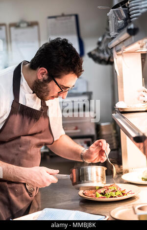 Bearded chef wearing glasses and apron standing in commercial kitchen at counter, plating food. Stock Photo