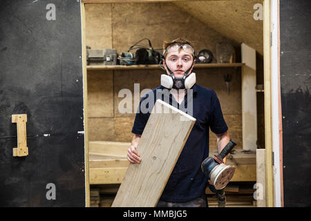 Young man wearing dust mask and protective goggles standing in a workshop, holding sander piece of wood. Stock Photo