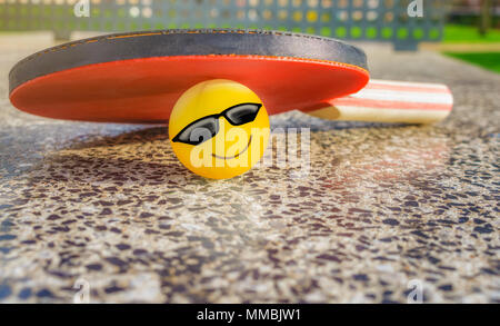 Tennis table paddle with a smiley ball below it on a stone tennis table in a park. Loving sport concept. Stock Photo