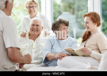 Smiling nurse holds book and sitting next to happy elder people in senior home Stock Photo