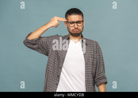 Portrait of bearded sad depressed man with black glasses in casual style looking at camera and holding hand with gun sign . studio shot on blue backgr Stock Photo