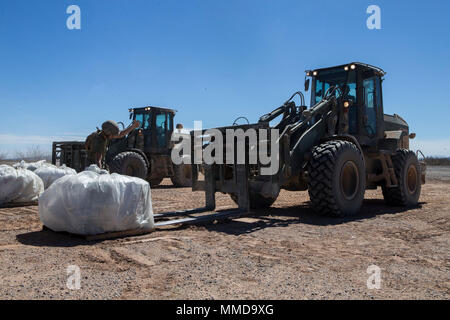 U.S. Marine Corps Pfc. Myles Bautista, a heavy equipment operator with Marine Wing Support Squadron 272 (MWSS-272) ground guides Pfc. Ashley Hewes, and Cpl. Timothy Lee, a heavy equipment operators with MWSS-272, while they lift bags of asphalt using a Tractor, Rubber Tired, Articulated, Multipurpose vehicle (TRAM) during expeditionary airfield maintenance in support of Weapons and Tactics Instructors course (WTI) 2-18 at STOVAL Expeditionary Airfield, Dateland, Ariz., March 14, 2018. WTI is a seven-week training event hosted by Marine Aviation Weapons and Tactics Squadron One (MAWTS-1) cadre, Stock Photo