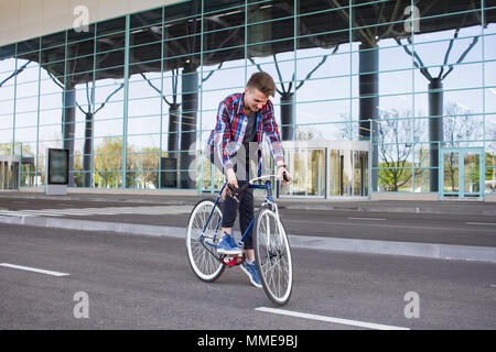 young man riding on bicycle in city street. Man on blue bicycle with white wheels, big mirror windows background Stock Photo