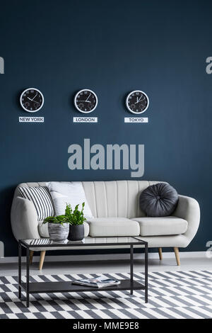 Clocks showing the hour in different time zones on a dark blue wall, above a comfortable beige sofa in a modern apartment interior Stock Photo