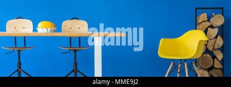 Creative plastic yellow chair by firewood in a stand and a dining table in a royal blue apartment interior Stock Photo