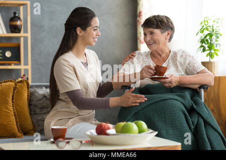 Young nurse serving coffee to senior lady in a wheelchair. Home visit concept. Stock Photo