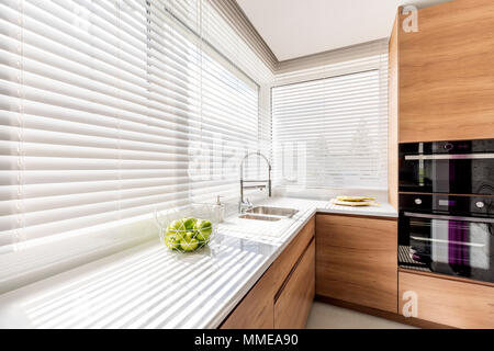 Modern bright kitchen interior with white horizontal window blinds, wooden cabinets with white countertop and household appliances Stock Photo