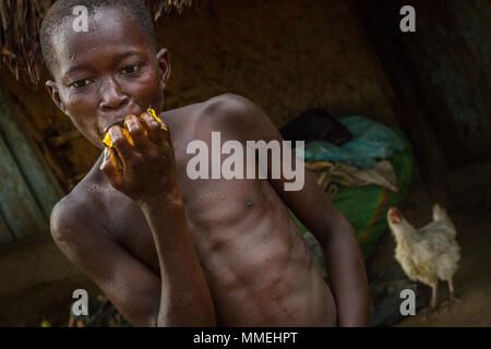 YONGORO, SIERRA LEONE - June 06, 2013: West Africa, portrait of an unknown boy while eating a yellow fruit in the background a chicken, near the capit Stock Photo