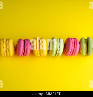 colored baked cakes of almond flour with cream on a yellow background, dessert macarons lies in a row in the middle Stock Photo