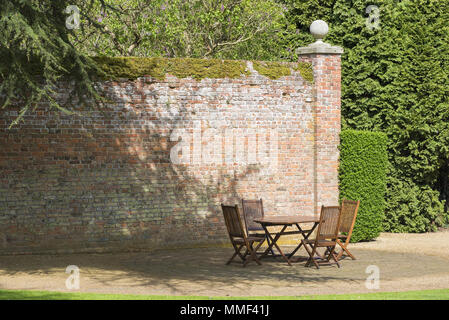Picinc table and chairs in a shady part of a garden surrounded by old red brick wall and tall green trees.