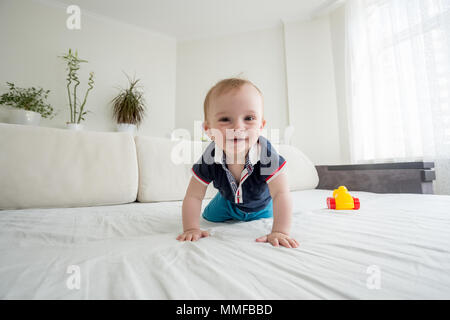 Wide angle photo of smiling toddler boy crawling on bed and looking in camera Stock Photo