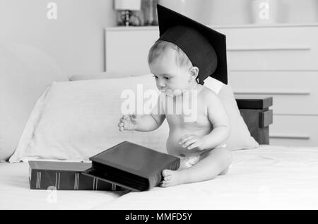 Black and white photo of baby boy in graduation cap sitting on bed and looking on big pile of books Stock Photo