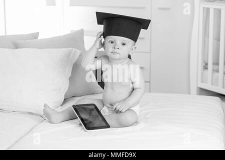 Black and white photo of little baby boy in graduation cap holding digital tablet Stock Photo