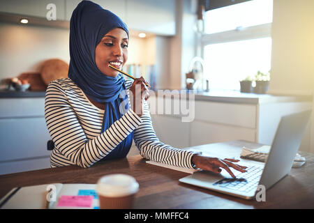 Smiling young Arabic female entrepreneur wearing a hijab sitting at her kitchen table working online with a laptop Stock Photo