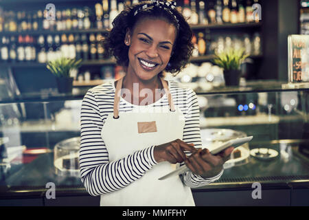 Smiling young African entrepreneur wearing an apron standing in her trendy cafe using a digital tablet Stock Photo