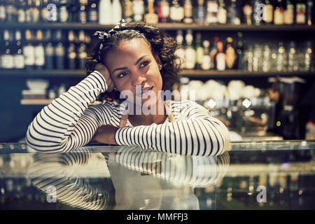 Young African cafe owner smiling and looking deep in thought while leaning on the counter of her trendy cafe