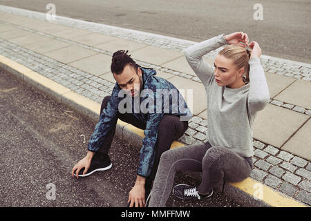 Diverse young couple in sportswear sitting by a road and catching their breath while jogging together outdoors Stock Photo