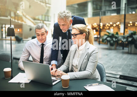 Three business colleagues talking together over a laptop while having a meeting at a table in the lobby of an office building Stock Photo