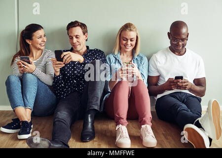 Smiling group of diverse young coworkers sitting together on the floor of an office using their cellphones during a break Stock Photo