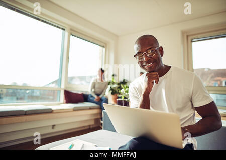 Smiling African businessman sitting on his office desk working on a laptop with coworkers sitting together in the background Stock Photo