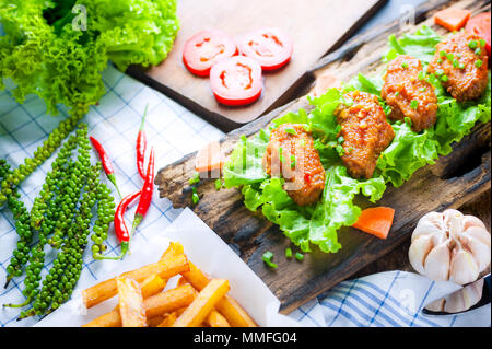 Fried chicken wings with spicy chilli sauce on rustic serving board, herbs and vegetable over wooden background. Stock Photo