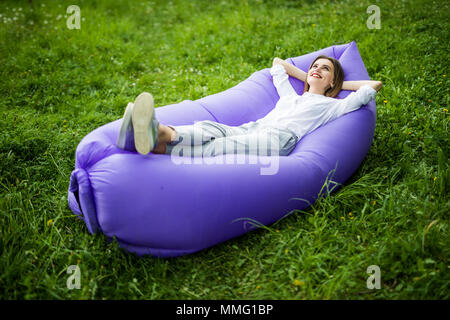 Pretty young woman lying on inflatable sofa lamzak while resting on grass in park Stock Photo