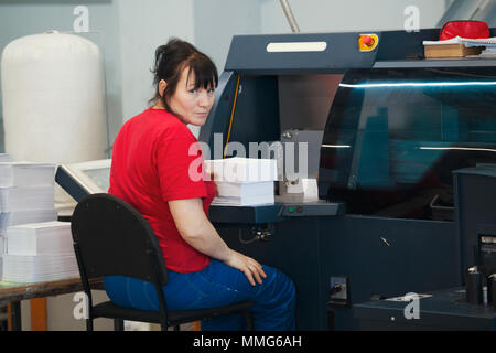 Adult female worker with a stacks of paper in front of print press equipment Stock Photo