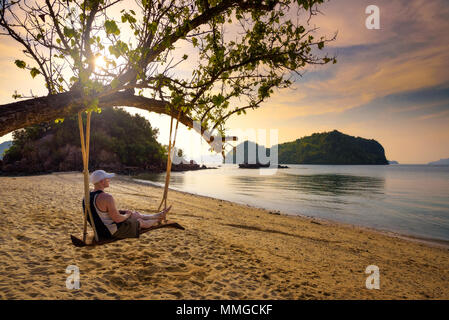 Young man enjoys sunset on a swing at a beach in Thailand Stock Photo