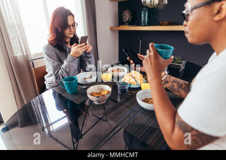 Couple using mobile phone while having morning breakfast at home. Man and woman sitting at breakfast table using smart phone. Stock Photo