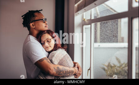 Interracial couple hugging each other while standing near window. African man embracing his caucasian girlfriend at home. Stock Photo