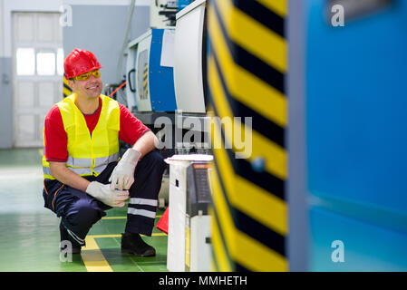 Worker in a factory. Worker wearing reflective west and red helmet crouching beside a machine Stock Photo