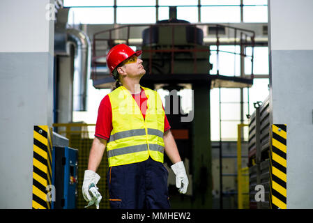 Worker in a factory looking up with machines in the background. Worker wearing reflective west and red helmet Stock Photo