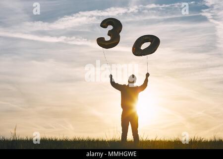 Man celebrates thirty years birthday. Person holding helium balloons in shape of number 30 at sunset. Stock Photo