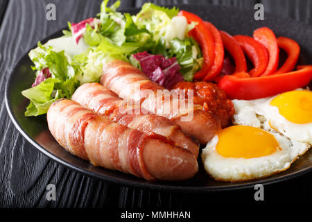 Breakfast Pigs in blankets fried sausages wrapped in bacon, eggs, sauce and fresh salad close-up on a plate. horizontal Stock Photo