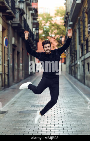 Young bearded man jumping in urban background with open arms wearing casual clothes. Guy with beard and modern hairstyle in the street. Stock Photo