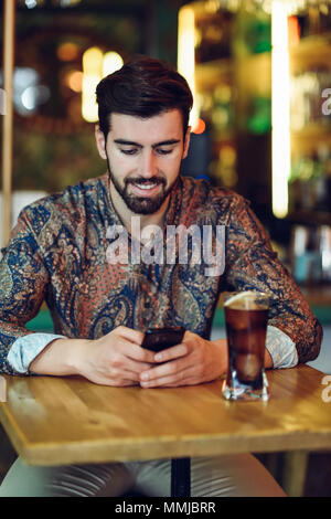 Young bearded man wearing casual clothes looking at his smartphone in a modern pub. Guy with beard and modern hairstyle drinking a cola. Stock Photo