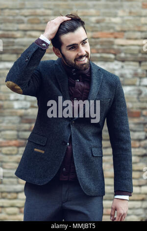 Young bearded man, model of fashion, smiling in urban background wearing british elegant suit. Guy with beard and modern hairstyle in the street. Stock Photo