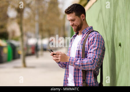 Smiling young man looking his smart phone in urban background. Guy wearing casual clothes. Lifestyle concept. Stock Photo