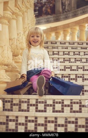 Blonde girl 3 years old in bright clothes sitting on the stairs with shopping Stock Photo