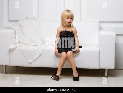Girl 3 years old in her mother's high heels sitting on a white sofa Stock Photo