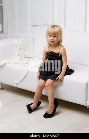 Girl 3 years old in her mother's high heels sitting on a white sofa Stock Photo