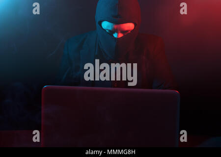 Angry computer hacker in suit stealing data from laptop illuminated by red and blue light Stock Photo