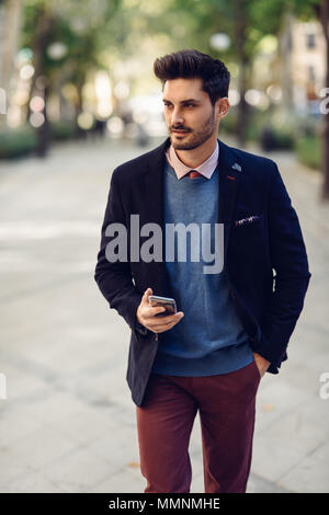 Attractive man in the street wearing british elegant suit with smart phone in his hand. Young bearded businessman with modern hairstyle in urban backg Stock Photo