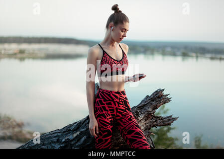 A beautiful brunette girl in stylish sports clothes is resting after a sport active workout near the lake. Listens to music on headphones with a smart Stock Photo